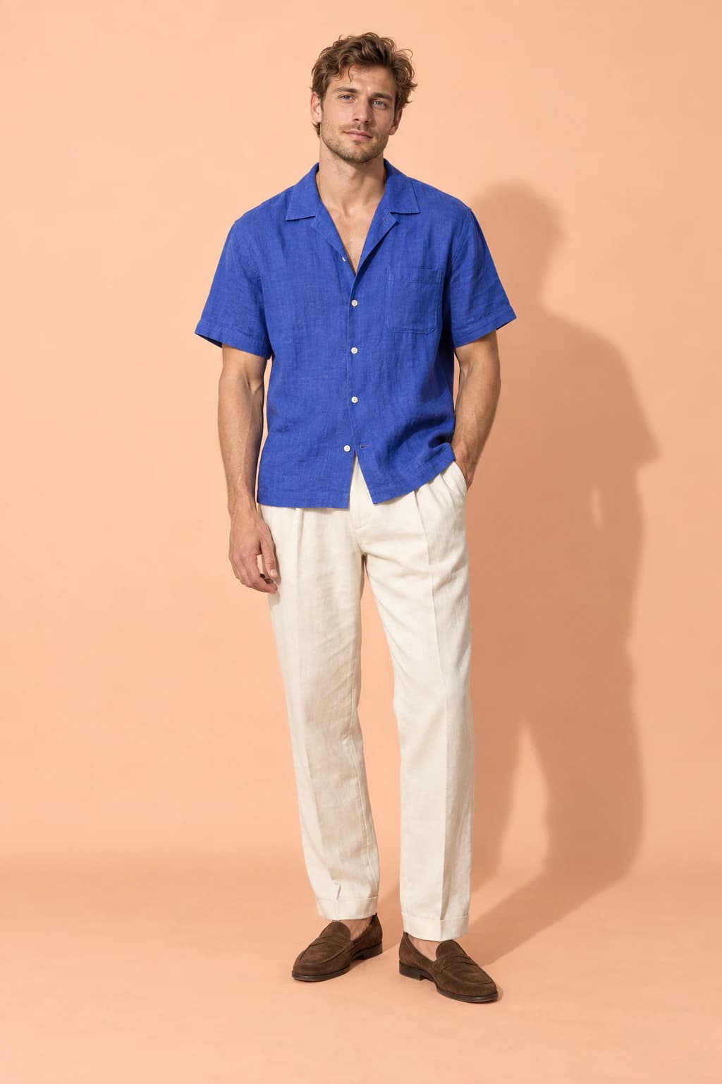 Male model in a cobalt linen camp-collar shirt and cream trousers on a peach backdrop