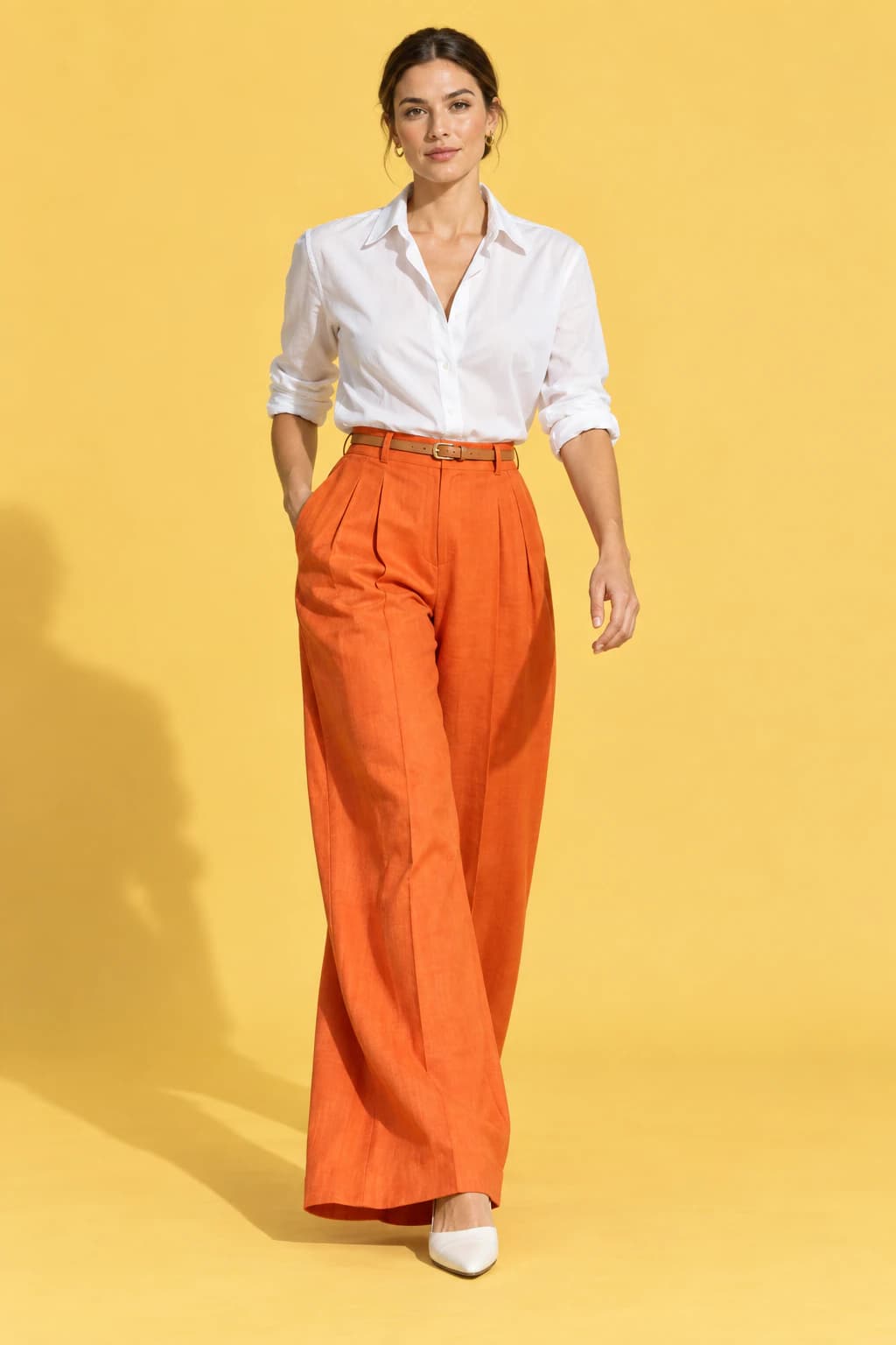 Model walking in tangerine wide-leg linen trousers and a white shirt on a butter-yellow backdrop